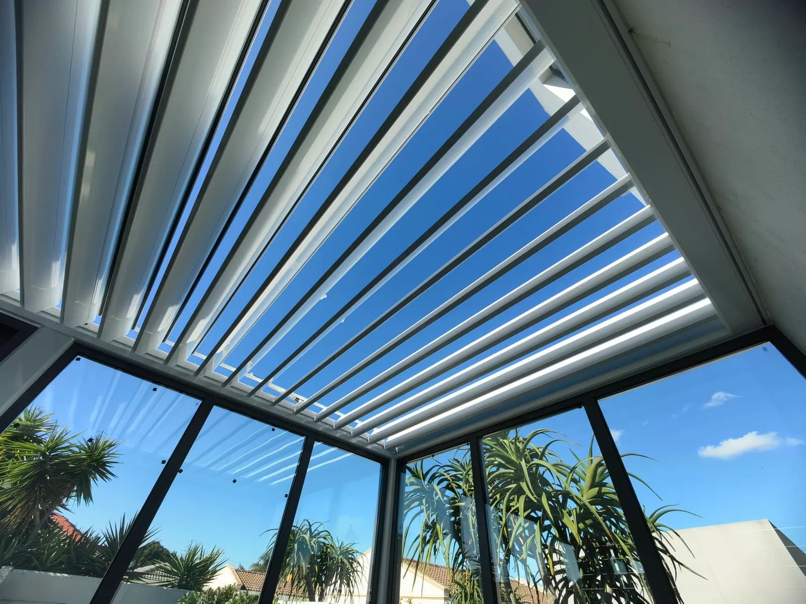 Looking up through open white louvres to a blue sky with palm trees beyond the glass walls
