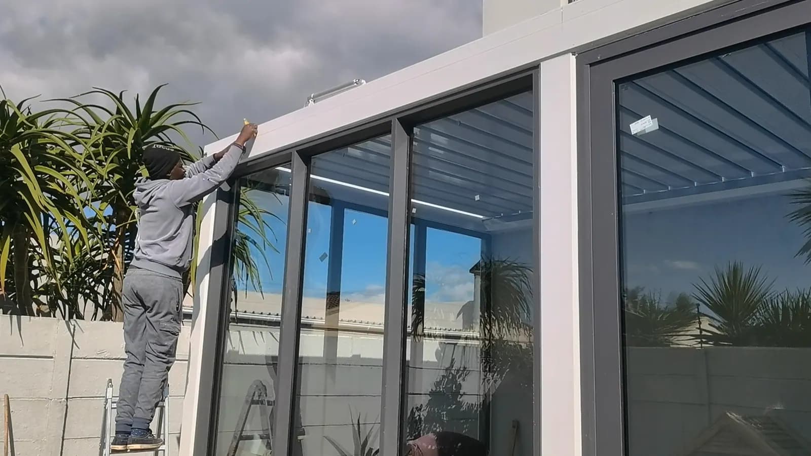 An installer on a ladder sealing the top of a glass wall panel under a louvred roof